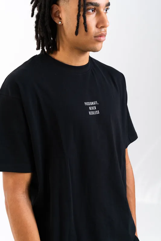 Young man with shoulder-length locs modelling a black printed T-shirt against a white wall