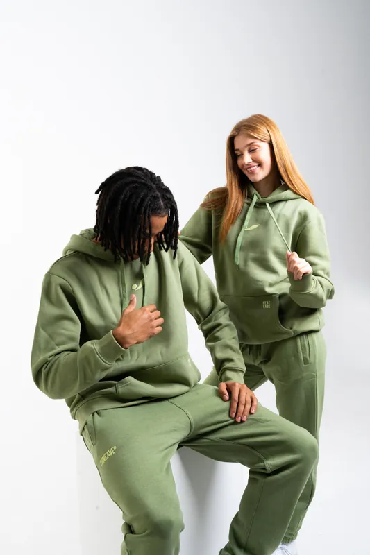 Two models in matching sage-green hoodies and track pants laughing together in a white studio