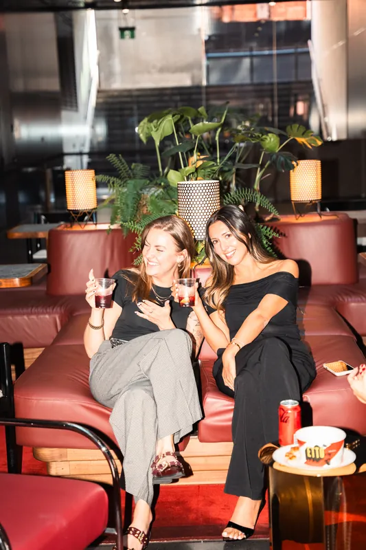 Two women laughing with drinks on a red leather booth in a warmly lit bar