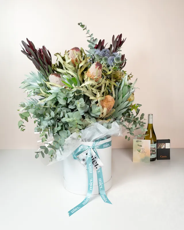 Native flower arrangement in a ribboned hat box beside a bottle of wine and a gift card