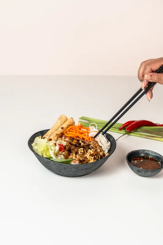 Chopsticks lifting noodles from a vermicelli salad bowl with spring rolls, chilli and a dipping sauce