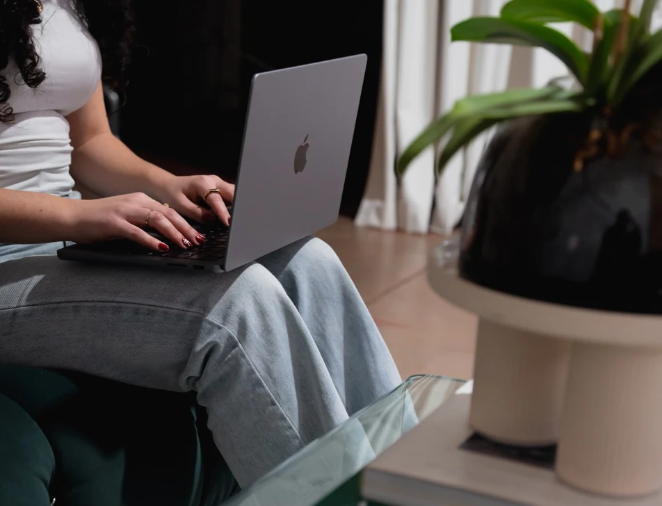 A woman typing on a laptop on her lap beside a glass coffee table and a potted plant
