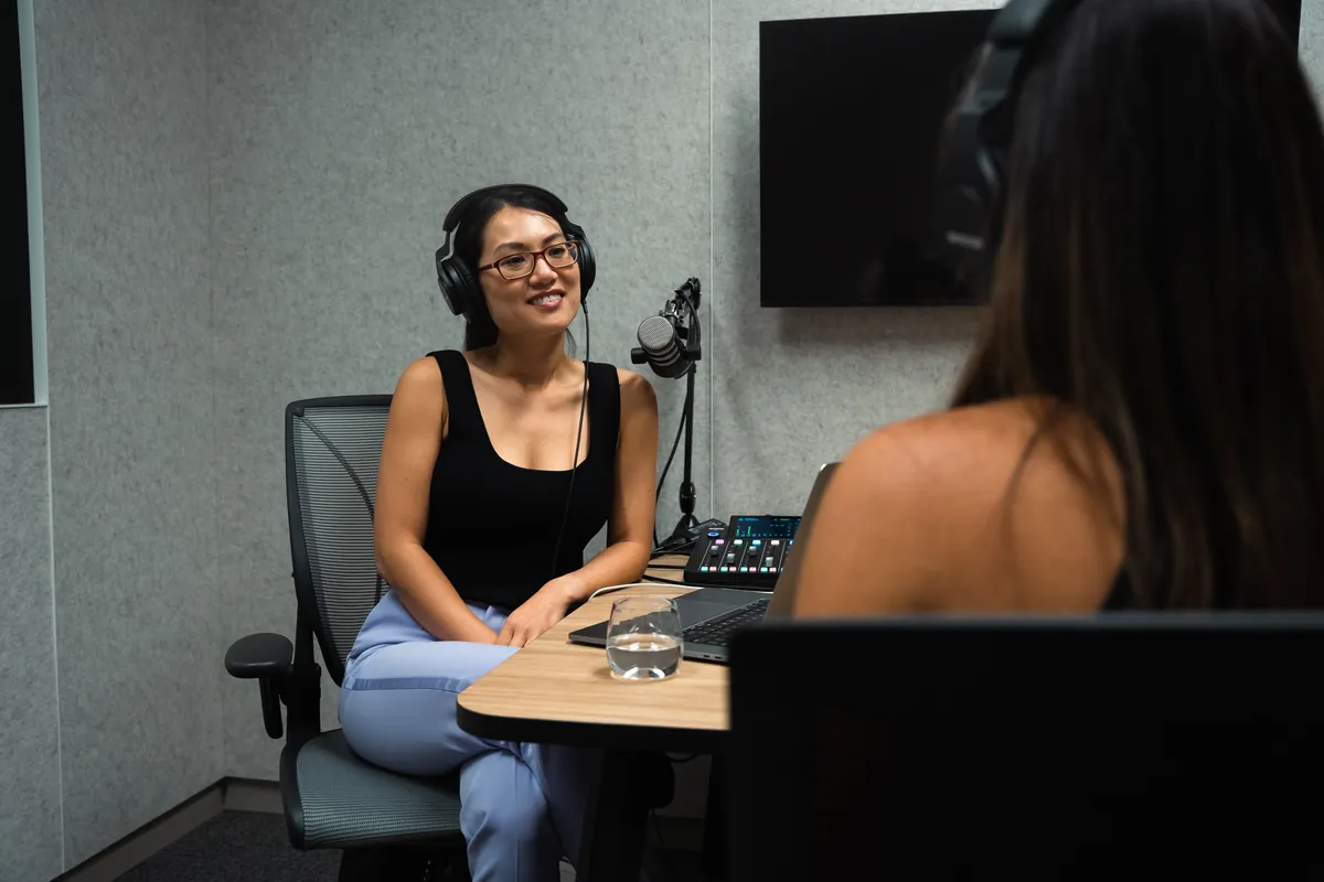 Woman in headphones smiling across a podcast studio desk at another person in headphones