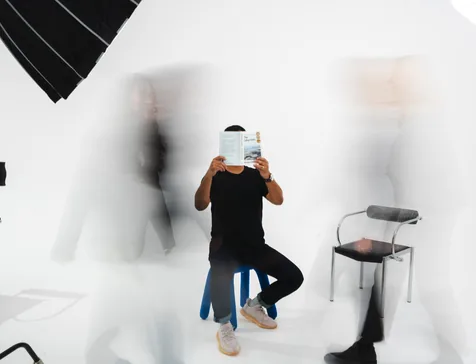 A person seated in a white studio holding a phone, with motion-blurred figures around them