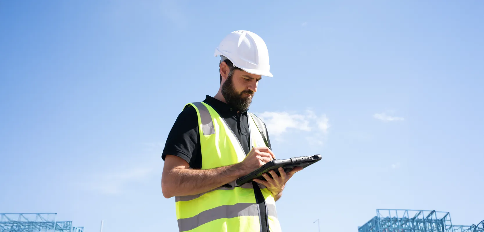 Site engineer in a white hard hat and hi-vis vest writing on a tablet against a clear blue sky