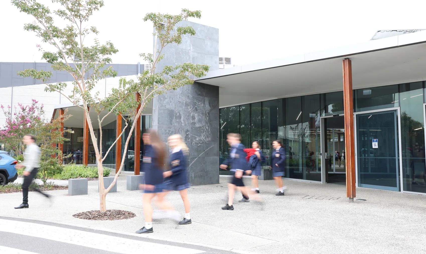 Students in St Francis Xavier College uniforms walking past the stone-and-glass entrance of a campus building