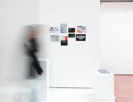 A person walking past printed work pinned to a white studio wall, blurred in motion
