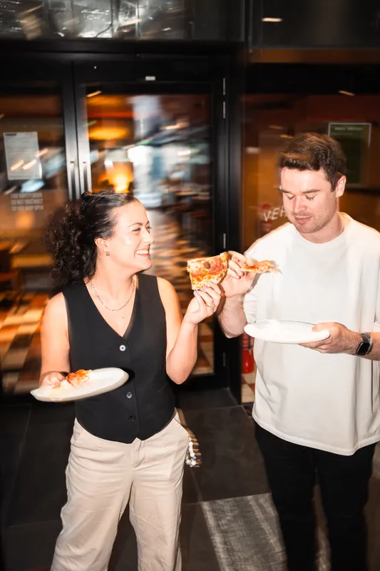 Two guests sharing slices of pizza inside a warmly lit venue
