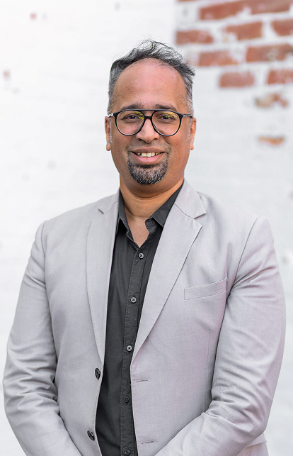 Portrait of Jobby Mathew, smiling in a gray shirt and suit against a white brick wall