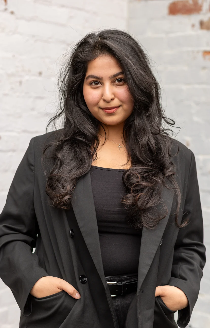 Portrait of Angel Makwana in a black blazer against a white brick wall