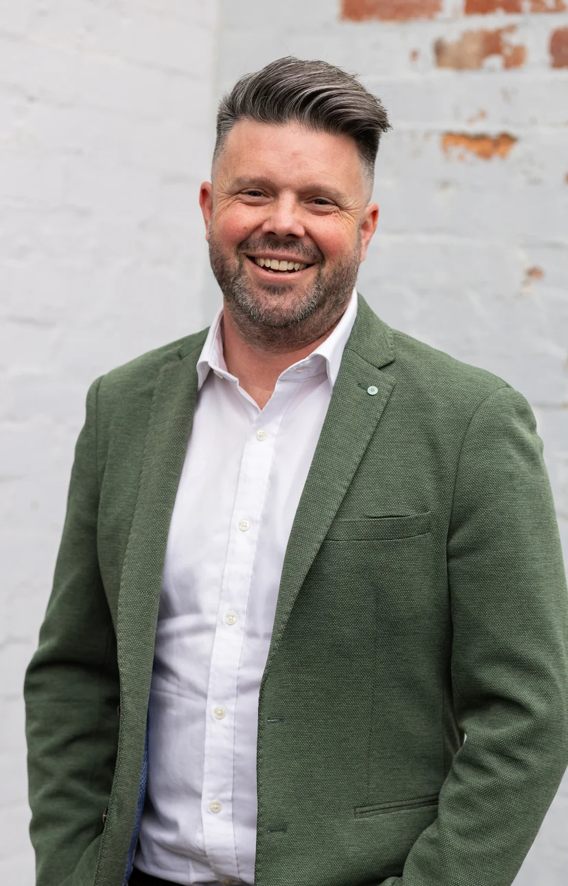 Portrait of Clint Chambers, smiling in a green blazer against a white brick wall
