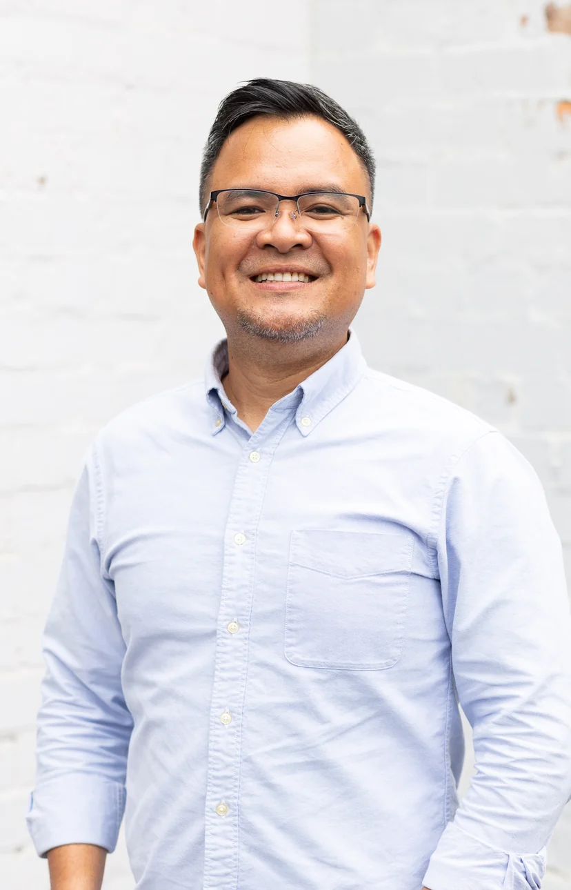 Portrait of Ian Molina, smiling in a pale blue shirt against a white brick wall