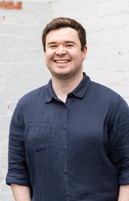Portrait of Lachlan Kempson, smiling in a navy shirt against a white brick wall