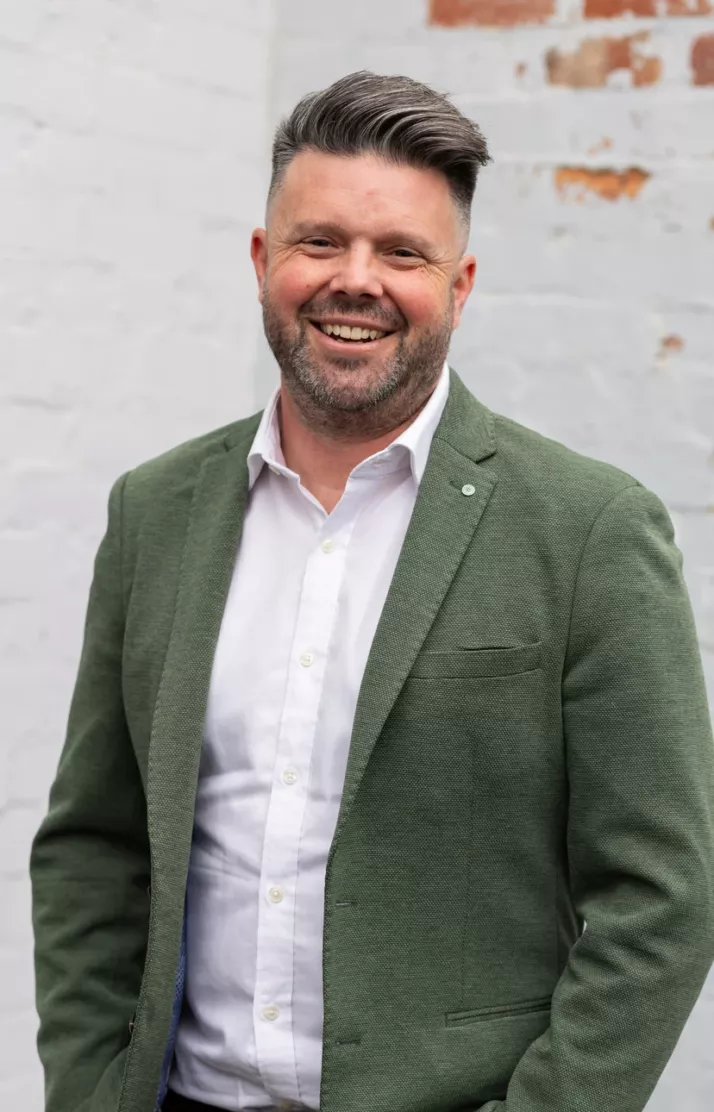 Portrait of Clint Chambers, smiling in a green blazer against a white brick wall