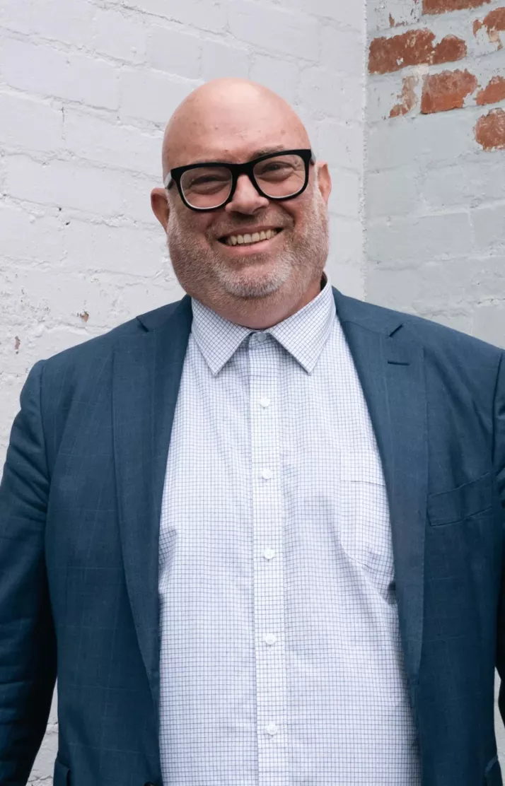 Portrait of Dave Hagger, smiling in a teal blazer and glasses against a white brick wall