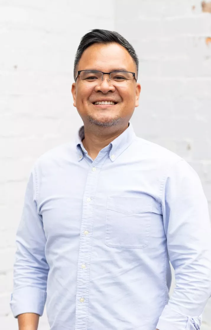 Portrait of Ian Molina, smiling in a pale blue shirt against a white brick wall
