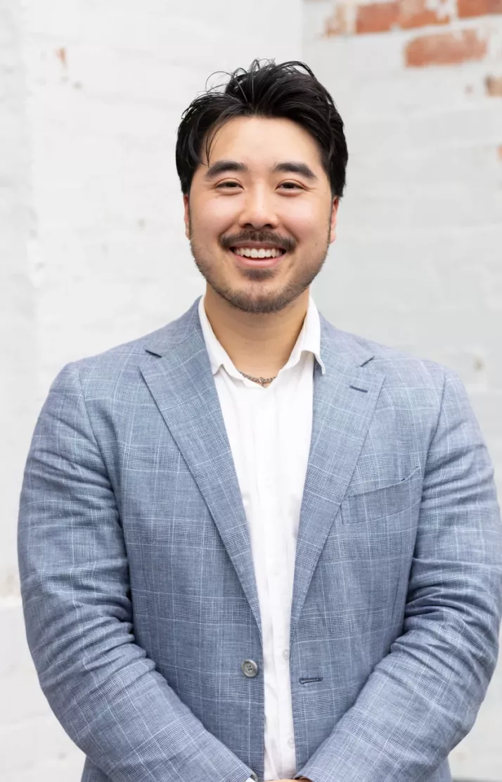 Portrait of Matthew Nguyen, smiling in a light blue blazer against a white brick wall